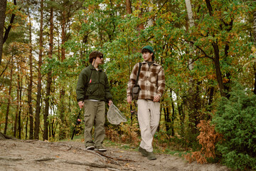 Naklejka premium Two friends walk through a vibrant autumn forest, preparing for a fishing trip. They carry fishing rods and a net as leaves fall around them, showcasing the beauty of nature.