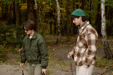 Fototapeta premium Two friends walk along a forested area, holding fishing rods. They are dressed in warm clothing, ready to enjoy their relaxing day by the water surrounded by colorful autumn leaves.