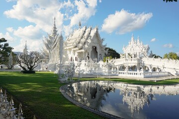 Majestic White Temple (Wat Rong Khun) in Chiang Rai, Thailand, featuring intricate architecture and a stunning mirror reflection on a calm pond under a clear blue sky.