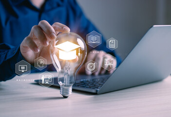 concept of online education courses or e-learning technology, man's hand touching a light bulb with graduation cap on desk, internet training on education, modern knowledge acquisition