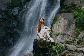 Obraz premium Young woman meditating peacefully on rock near waterfall surrounded by lush green nature and moss-covered stones, wearing white activewear in serene outdoor setting.