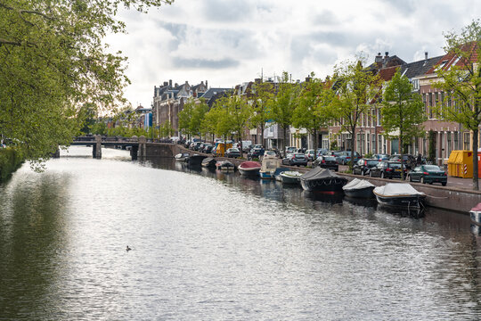 View of the tranquil canal reflecting the sky, lined with parked cars, boats, and charming buildings under a soft, overcast sky, Haarlem, North Holland, Netherlands. - Powered by Adobe