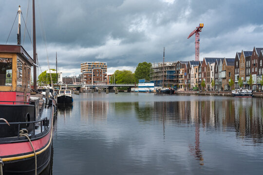 View of calm canal waters reflecting brick buildings and a construction crane under a cloudy sky, a boat with red trim in the foreground, Haarlem, North Holland, Netherlands.