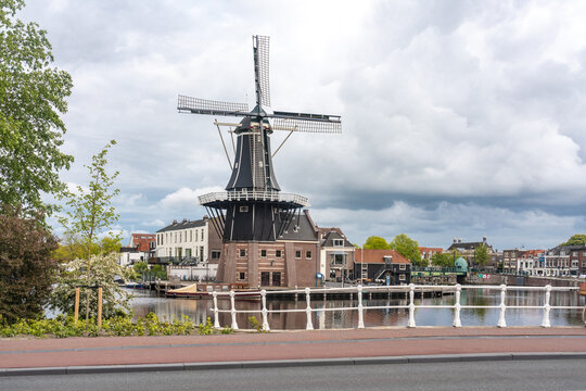 View of a historic windmill standing tall beside a canal under a cloudy sky, its dark wood contrasting with the light buildings, Haarlem, North Holland, Netherlands.