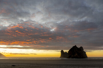View of dramatic skies ablaze with fiery hues meet the dark silhouette of coastal rock formations on a serene beach, Wharariki Beach, Pūponga, Tasman Region, New Zealand.