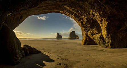 View of golden sands stretching toward the horizon, framed by the rugged mouth of a dark cave, revealing two towering rock formations, Wharariki beach, Pūponga, Tasman Region, New Zealand.