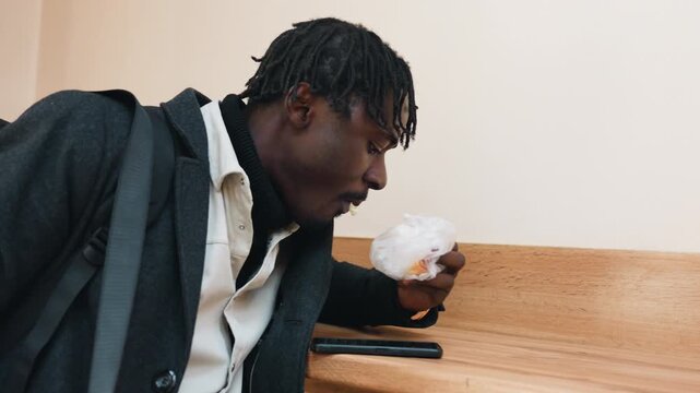 African man munches dough ball while leaning hand on table, dressed in dark coat and light shirt, showing natural gesture and thoughtful expression in warm indoor setting with relaxed everyday mood