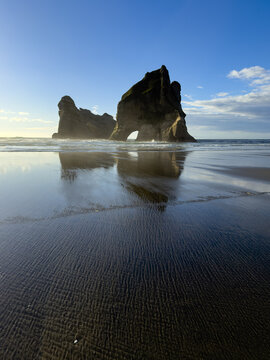 View of majestic rock formations rise from the dark, wet sand, mirrored in the receding tide under a vibrant blue sky, Wharariki beach, Pūponga, Tasman Region, New Zealand.