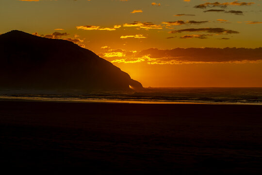 View of sun ablaze, golden light kisses the ocean and silhouettes the rugged coastline in a dance of shadow and fire, Pūponga, Tasman Region, New Zealand.