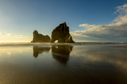 View of colossal rock formations reflected on the wet sandy Wharariki beach under a blue sky touched with soft clouds, Pūponga, Tasman Region, New Zealand.
