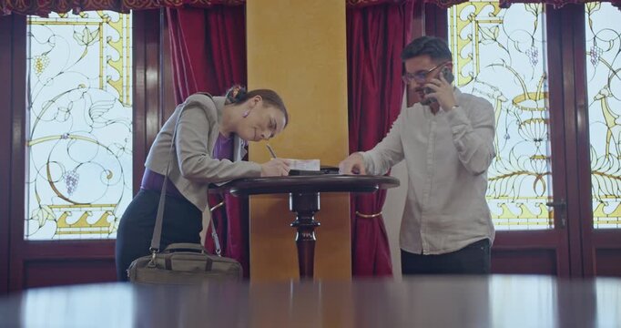 Business partners discuss documents in a hotel lobby, one taking a phone call. They are formally dressed and focused on their tasks.
