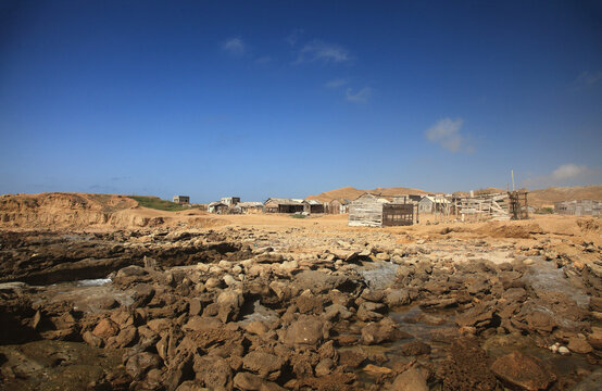 View of rocky terrain leading to modest buildings under a vast blue sky, capturing the raw beauty of a humble settlement, Karachi, Sindh, Pakistan.
