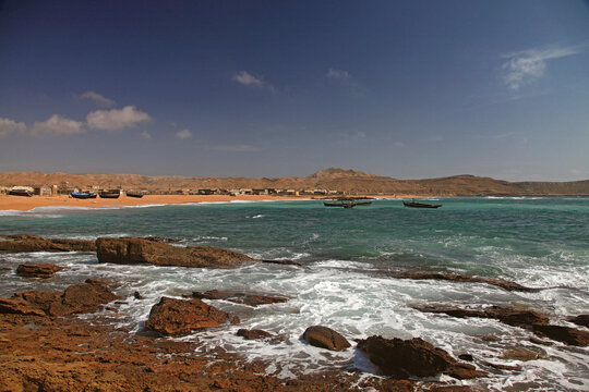 View of turquoise waves crashing on a rocky shore, contrasting with the sandy beach and distant structures under a clear sky, Karachi, Sindh, Pakistan.