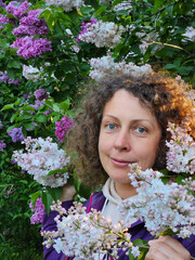 Woman is holding a bunch of white flowers. She is wearing a purple jacket in lilac park