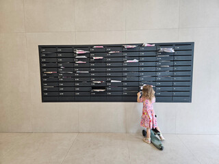 Little girl with scooter is standing in front of a wall with a black post box near entrance in the courtyard of the house in residential complex