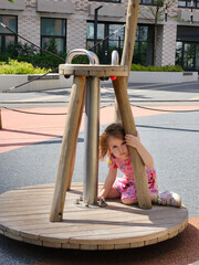 Little girl is sitting and thinking on a wooden merry go round. She is wearing a pink dress. The merry go round is made of wood and has a metal pole in the middle