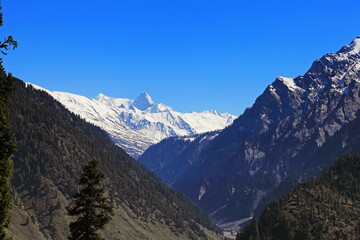 View of towering, snow-capped mountains pierce the azure sky, embraced by lush, verdant valleys cloaked in dense forest, Neelum Valley, Azad Kashmir, Pakistan.