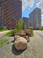 Girl baby is sitting on a bench in front of a large rock. The rock is surrounded by other rocks near entrance in the courtyard of the house in residential complex
