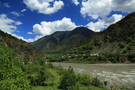 View of a rushing river snaking through a lush green valley beneath towering mountains under a bright blue sky with fluffy white clouds, Neelum Valley, Azad Kashmir, Pakistan.