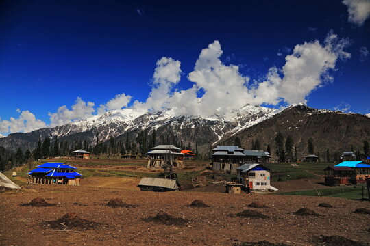 View of houses dot a landscape of brown fields, framed by snow-capped mountains under a vibrant blue sky, creating a stark contrast of color and texture, Neelum Valley, Azad Kashmir, Pakistan.
