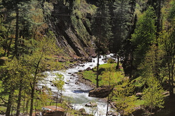 View of a rushing river carving its way through a steep, verdant valley, flanked by dense forests and rocky slopes in the heart of nature, Neelum Valley, Azad Kashmir, Pakistan.