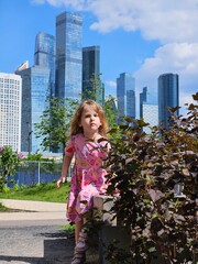 Little girl in a pink dress is standing in front of a bush. The bush is green and has some brown leaves on skyscrapers