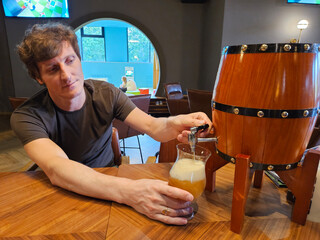 Man is pouring beer from a barrel into a glass. The barrel is wooden and has a metal handle. Playroom for children in restaurant