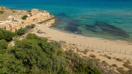 Aerial view of a wild beach overlooking a crystalline sea. The sea water hues range from turquoise to blue. There is no one on the sand and the shore is empty.