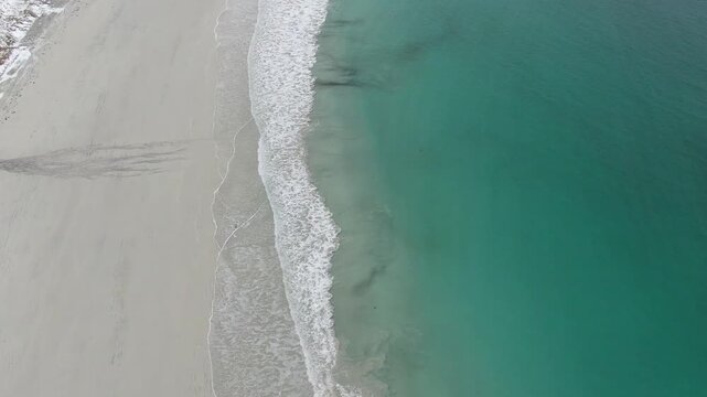 A drone view over Ramberg Beach in the Lofoten Islands reveals icy winter scenery, with snow-covered shores and striking turquoise-blue water under cold Arctic light.