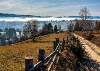 Countryside Mountain Road with Wooden Fence in Rural Landscape. Sunshine in the late autumn mountain landscape with a fence and dirt road above misty valleys, Harghita County, Livezi village, Romania.
