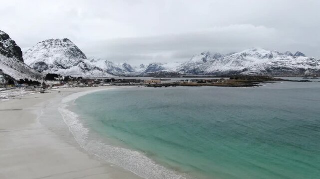 A drone view over Ramberg Beach in the Lofoten Islands reveals icy winter scenery, with snow-covered shores and striking turquoise-blue water under cold Arctic light.