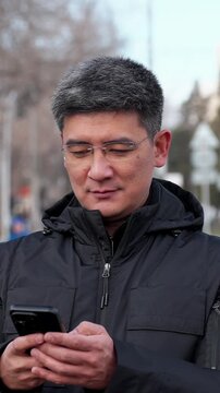 Middle-aged asian man in glasses using smartphone outdoors on city street. Vertical shot of mobile communication, digital lifestyle, online services, messaging and modern urban technology