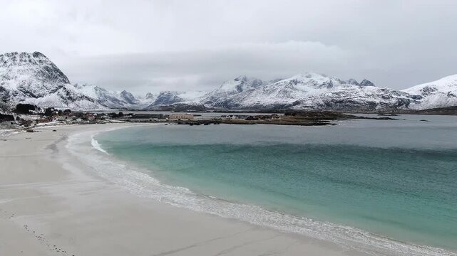 A drone view over Ramberg Beach in the Lofoten Islands reveals icy winter scenery, with snow-covered shores and striking turquoise-blue water under cold Arctic light.
