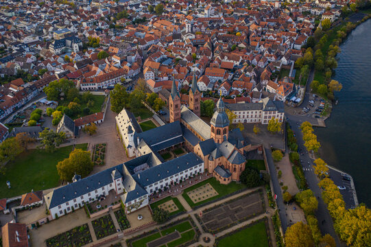 Aerial view of the Einhard Basilica's dark stone contrasts against the bright, warm hues of the surrounding rooftops and the Main river, Seligenstadt, Hessen, Germany.