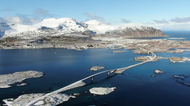 A drone view of the bridges to Fredvang in the Lofoten Islands on a sunny winter day, with snow-covered mountains and crystal-clear water creating a true Arctic paradise.