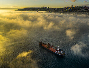 Aerial view of a lone cargo ship sailing through the misty waters near Haifa, with the city's skyline faintly visible, Haifa, Israel.