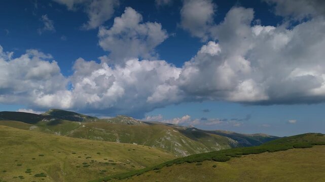 Bucegi Mountains aerial panorama over the high alpine plateau, revealing Scara, Omu, Gavanele, Costila and Piatra Arsa peaks beneath dramatic fast-moving summer clouds and deep blue sky, Romania