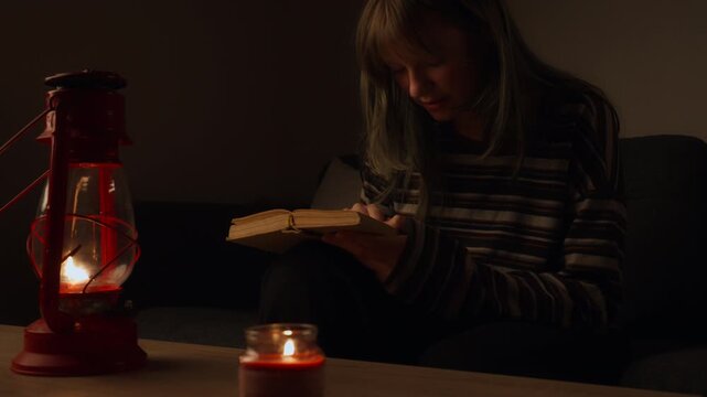 Young woman sitting on sofa and reading an old book in dark room, illuminated only by warm, flickering light of red kerosene lantern and single candle during power outage