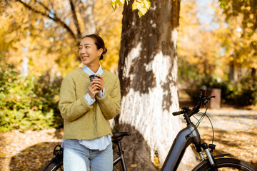 Korean woman enjoying warm beverage while standing by bicycle in autumn park