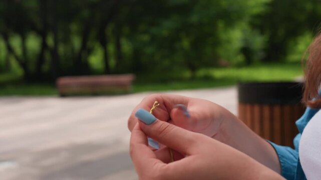 Close up hand view of person separating green thread with blurred park background, blue nails and steady fingers preparing embroidery outdoors, calm focus untangling floss strand under soft daylight