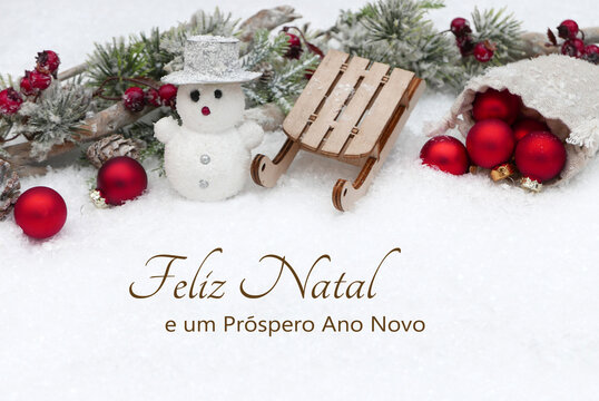 Cart&atilde;o de Natal com votos de Ano Novo em portugu&ecirc;s: Bolas de Natal, um boneco de neve com um tren&oacute; e ramos de pinheiro na neve. "Feliz Natal e um Pr&oacute;spero Ano Novo".