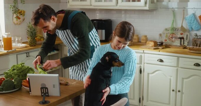 Father watches a recipe tutorial on a tablet while chopping vegetables in the kitchen. Daughter plays with the family dog nearby. Warm family atmosphere and everyday home life.