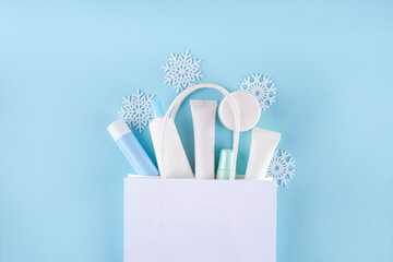 Winter skin care and hand care cosmetics sale and shopping, various beauty care cosmetic containers, bottles, tubes with artificial snowflakes and paper shopping bag on light blue background top view 