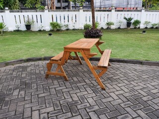 side angle view of a wooden foldable picnic table and bench set located on a brick patio. The background features a green lawn, white garden wall, and lush tropical trees.
