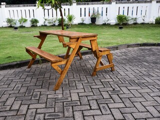 a wooden picnic table with attached benches stands on a brick-paved patio in a lush green garden. A white perimeter fence and small trees are visible in the bright background.