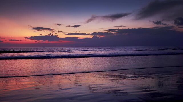 Gorgeous sunset landscape with seashore washed by waves of calm sea and silhouettes of people swimming in water against clouds lit by setting sun on background. Ocean beach in twilight. Still camera.