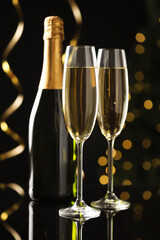 Merry Christmas and Happy New Year. Flute glasses with sparkling wine, serpentine streamers and bottle on black background against blurred lights