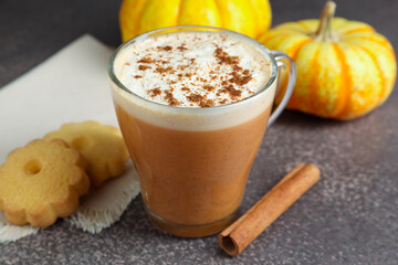 Delicious pumpkin latte in glass cup, cinnamon stick, cookies and fresh vegetables on grey textured table, closeup