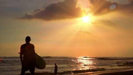 Unrecognizable figure of young slim male surfer holding surfboard and passing by on sand beach washed by sea waves and illuminated by evening sun. Bali, Indonesia. Slow motion. Camera zooms out.