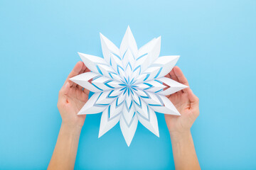 Woman hands holding and showing white snowflake shape from paper on light blue table background. Pastel color. Making decoration element for winter festive. Closeup. Point of view shot. Top down view.